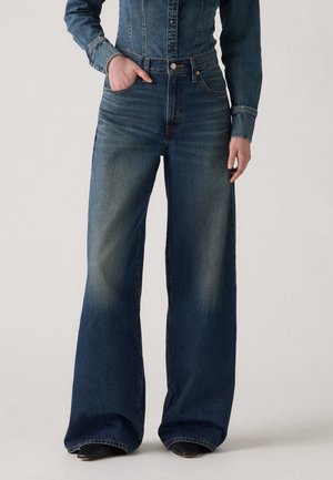 Personne portant un jean en denim bleu taille haute à jambes larges, main dans la poche, avec une veste en denim assortie, debout sur un fond clair.