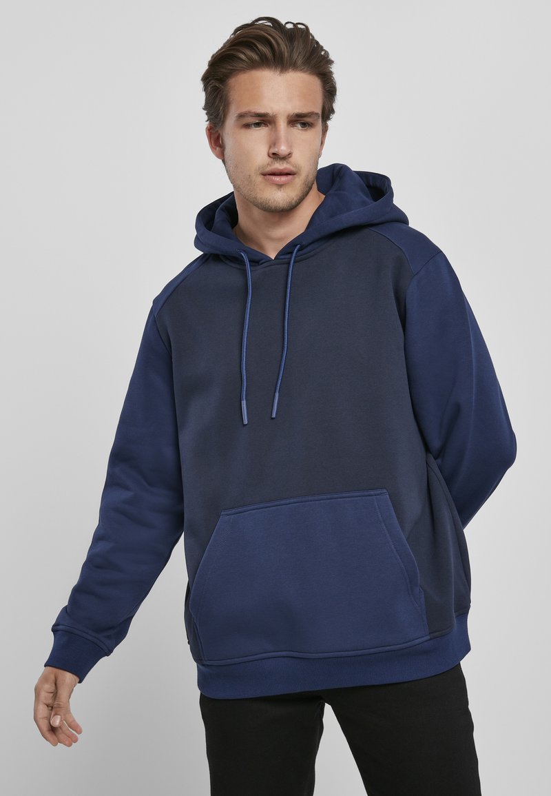 Felpa con cappuccio blu navy che presenta una tasca frontale, stringhe di regolazione e maniche in contrasto, realizzata in un tessuto morbido con una superficie liscia.