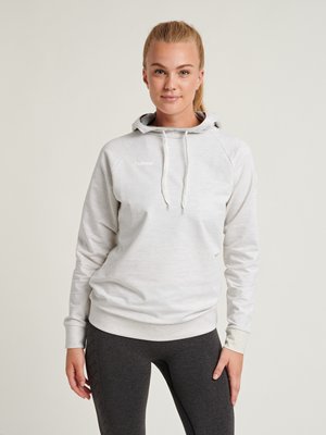 Kvinde med blondt hår i en hestehale iført en lysegrå hættetrøje og mørkegrå leggings, stående mod en ensfarvet neutral baggrund.