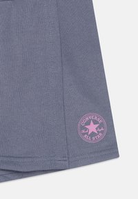 T-shirt en coton gris avec un logo rose rond représentant une étoile et le texte "Converse All Star" près de l'ourlet. Texture lisse, design décontracté.