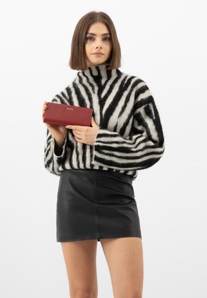 Femme aux cheveux bruns coupés au niveau du menton, portant un pull rayé noir et blanc et une mini-jupe en cuir noire, tenant un portefeuille rouge.