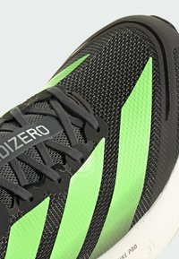 Chaussure de sport noire avec texture en maille, dotée de bandes vertes audacieuses. Comprend des lacets noirs et un logo "Adizero" sur le côté.