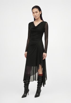 Femme portant une robe noire asymétrique avec des manches longues transparentes et des bottes noires à talons souples sur un fond blanc uni.