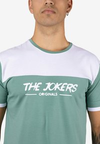 Camiseta de manga corta verde y blanca con un estampado gráfico "THE JOKERS ORIGINALS" en texto blanco en negrita en el pecho. Material de algodón.