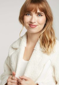 Veste en tissu blanc moelleux avec un col châle et deux poches avant. Superposée sur un haut blanc. Mains tenant les revers de la veste.
