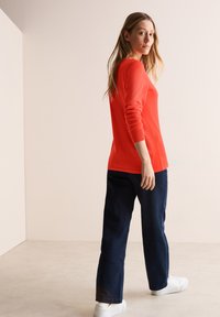 Rotes langärmliges Oberteil, kombiniert mit dunkelblauen Hosen und weißen Turnschuhen. Das Oberteil hat eine taillierte Form, gerippte Bündchen und eine glatte Textur.