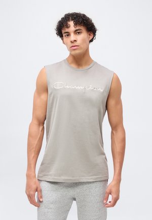 Ung mand med krøllet hår iført en ærmeløs grå Champion-shirt og lysegrå bukser, stående foran en ensfarvet baggrund.