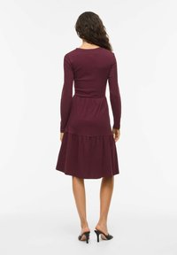 Robe longue à manches longues couleur bordeaux avec un corsage ajusté et une jupe évasée. Texture côtelée, encolure ronde et design à étages, portée avec des talons noirs.