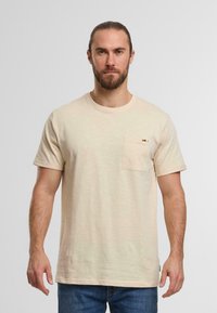 Light beige cotton t-shirt with a round neckline, short sleeves, and a front chest pocket. Subtle textured finish and casual fit.