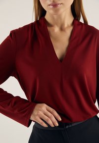 Červená blůza s dlouhými rukávy a hlubokým výstřihem do V, vyrobená z hladké látky, s jemným řasením na ramenou a elegantním střihem.