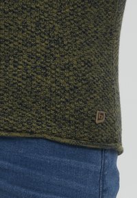 Grønn og marineblå teksturert strikkegenser med en brun stofflogo på undersiden, vist sammen med blå denim.