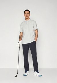 Světle šedé polotriko se třemi knoflíky a logem, kombinované s tmavě námořnickými kalhotami. Model drží golfovou hůl a má na sobě bílé tenisky.