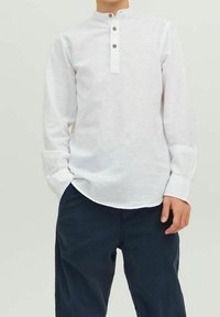 Biała koszula z długim rękawem i kołnierzykiem mandaryńskim, z trzema brązowymi guzikami, zestawiona z ciemnoniebieskimi spodniami o luźnym kroju i miękkiej fakturze.