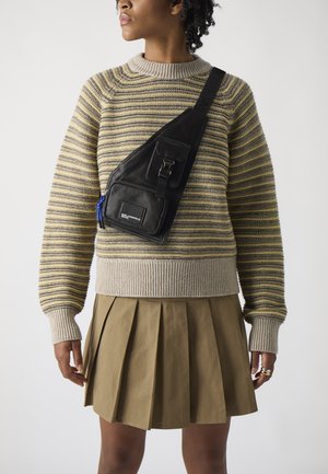 Black sling bag made of textured leather, with a front pocket, buckle detail, and blue zipper. Worn over a striped sweater and pleated skirt.
