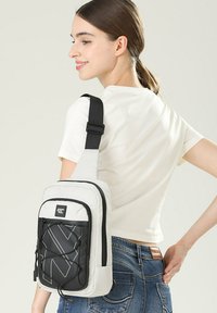 Borsa a tracolla grigia chiara con accenti neri, dotata di un pannello anteriore testurizzato e cinturino regolabile. La chiusura con cerniera e il dettaglio elastico ne aumentano la funzionalità.