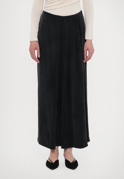 Jupe maxi plissée noire en tissu doux, avec une texture lisse et une forme légèrement évasée. Portée avec des ballerines noires à bout pointu.