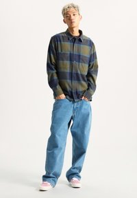 Camicia di flanella a quadri nei toni del verde, blu navy e blu, con due tasche sul petto, abbinata a jeans larghi di un blu chiaro e sneakers rosa.