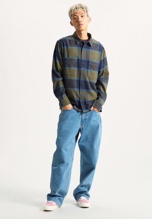 Camisa de franela a cuadros en tonos verdes, marinos y azules con dos bolsillos en el pecho, combinada con unos vaqueros sueltos de color azul claro y zapatillas rosas.