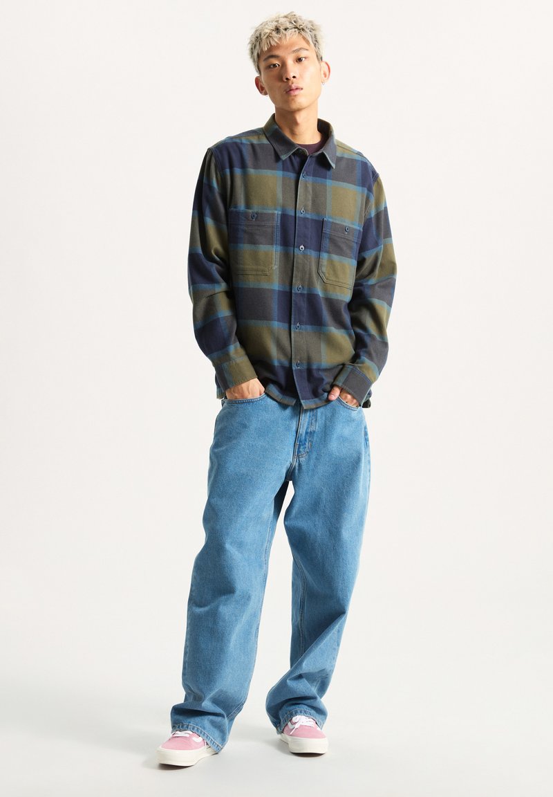 Camicia di flanella a quadri nei toni del verde, blu navy e blu, con due tasche sul petto, abbinata a jeans larghi di un blu chiaro e sneakers rosa.