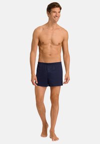 Námořnické modré bavlněné trenýrky s elastickým pasem a detaily knoflíku, model stojící bosý, předvádějící uvolněný střih a design.