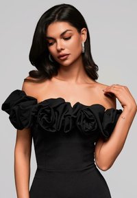 Robe noire épaules dégagées avec de grandes manches volantées, tissu lisse, silhouette cintrée et léger éclat. Les détails élégants mettent en valeur le décolleté.