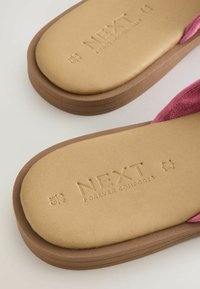 Gornji dio japanki od ružičastog antilopa s donjom stranom od smeškanog smeđeg gume. Ugravirani logo "NEXT FOREVER COMFORT" na bež ulošku. Ravni dizajn, ležeran stil.