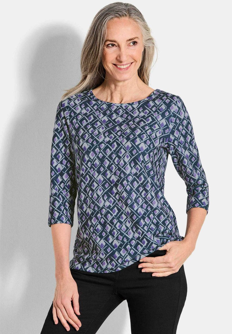 Langemmouwtop in tinten blauw en paars, met een geometrisch patroon, ronde halslijn en zachte textuur. Gecombineerd met zwarte broek.