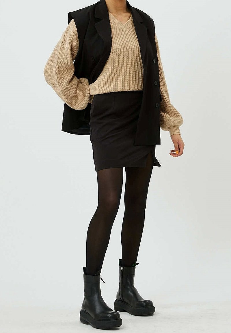 Personne portant un pull en maille beige avec des manches bouffantes, un blazer noir sans manches, une mini-jupe noire, des collants noirs et des bottines noires.