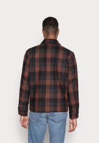 Brązowa i czarna kurtka w kratę z kołnierzem, mankietami na guziki oraz luźnym krojem, wykonana z miękkiego, teksturowanego materiału. Poniżej widoczne niebieskie dżinsy.