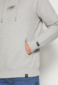 Sweat à capuche gris avec poche avant et patchs de logo noirs sur le poignet et l'ourlet, porté par une personne avec la main dans la poche, assorti à un jean bleu.