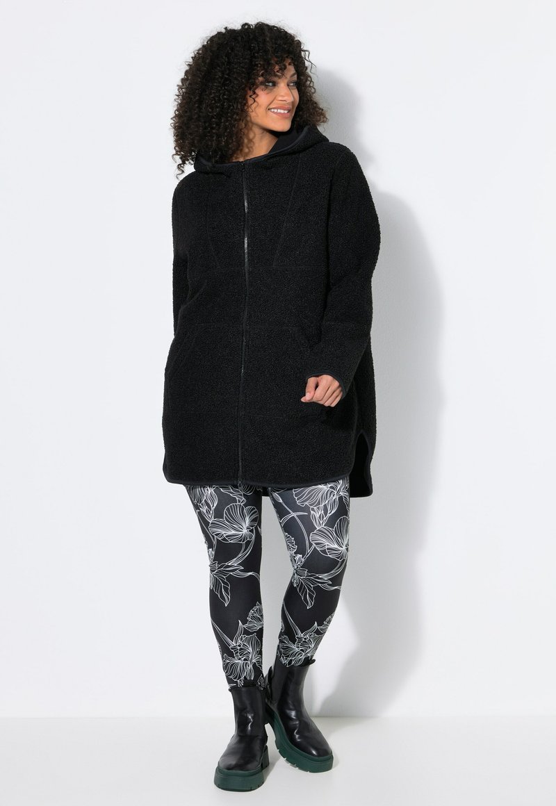 Mujer con cabello rizado que lleva una chaqueta de forro polar negra con capucha y cremallera, leggings florales en blanco y negro, y botas negras con suelas verdes.