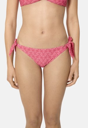Mujer con bragas de bikini con estampado floral rosa y lazos laterales, sobre un fondo liso y claro.