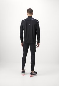 Tenue de sport noire comprenant un haut à manches longues et des leggings ajustés, avec des accents réfléchissants et une texture lisse et légère.