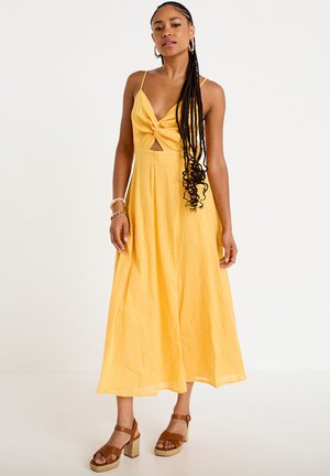 Femme aux longues tresses portant une robe midi jaune sans manches avec un nœud devant et des sandales marron à talons, debout sur un fond blanc.