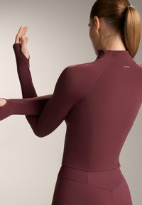 Bordó színű, testhezálló sportfelső hosszú ujjakkal, magas nyakkal és hüvelykujjszorítóval, rugalmas anyagból készült, apró logó részletekkel.