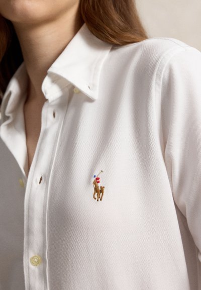 White collared shirt with an embroidered polo player logo with blue and red details on the chest, worn by a person with brown hair.