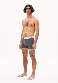 Männliche graue Boxer Shorts mit einem weißen elastischen Bund, der schwarzen Text trägt. Das Modell steht barfuß und zeigt Tattoos an den Beinen.
