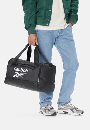 Osoba ubrana w jasnoniebieskie dżinsy, szaro-białe trampki i zieloną kurtkę, trzymająca czarną torbę sportową Reebok za uchwyty.