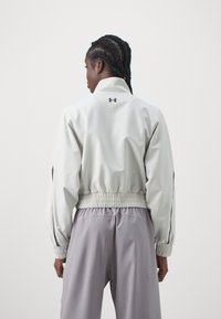 Light gray windbreaker jacket with a high collar, elastic waistband, and black accents. Features logo on the back and ribbed cuffs.