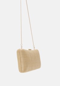 Pochette rectangulaire tissée beige avec bandoulière et fermoir en chaîne dorée, suspendue devant un fond blanc.