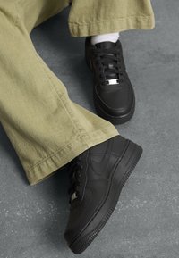 Svarte lær Nike Air Force 1-sneakers med perforert tå, flate skolisser og sølvfargede detaljer, brukt sammen med lysegrønne vide bukser.