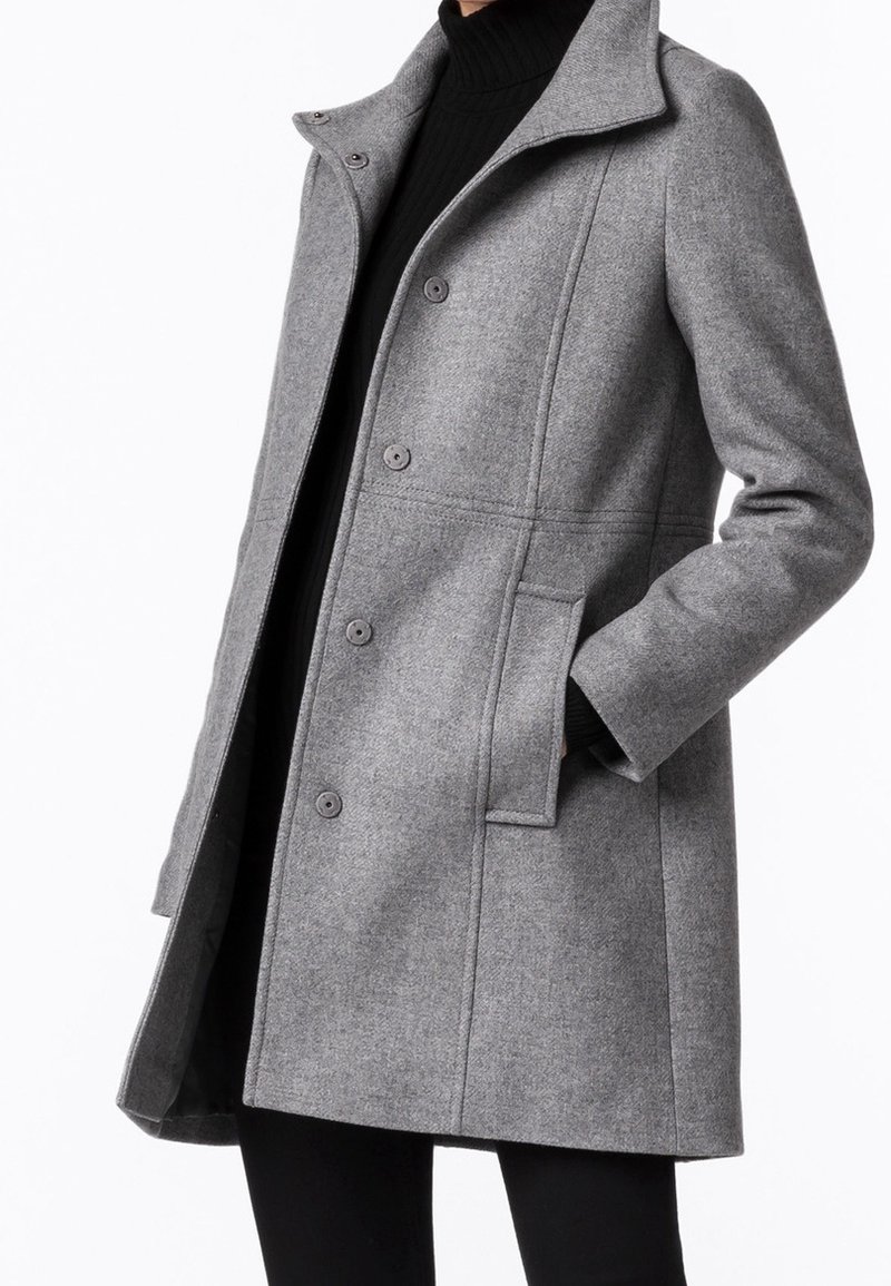Femme portant un manteau en laine gris mi-long avec boutons et poches latérales sur un col roulé noir et un pantalon, debout avec une main dans la poche.