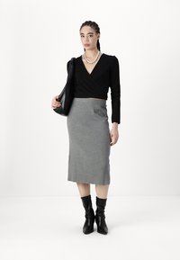 Top negro de manga larga, falda lápiz gris con relieve y botas negras hasta la rodilla. La modelo sostiene un bolso negro, de pie sobre un fondo blanco.