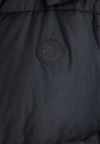 Black puffer jacket fabric with a smooth texture; features a circular black rubber logo with "HGO" and a smiley design at the center.