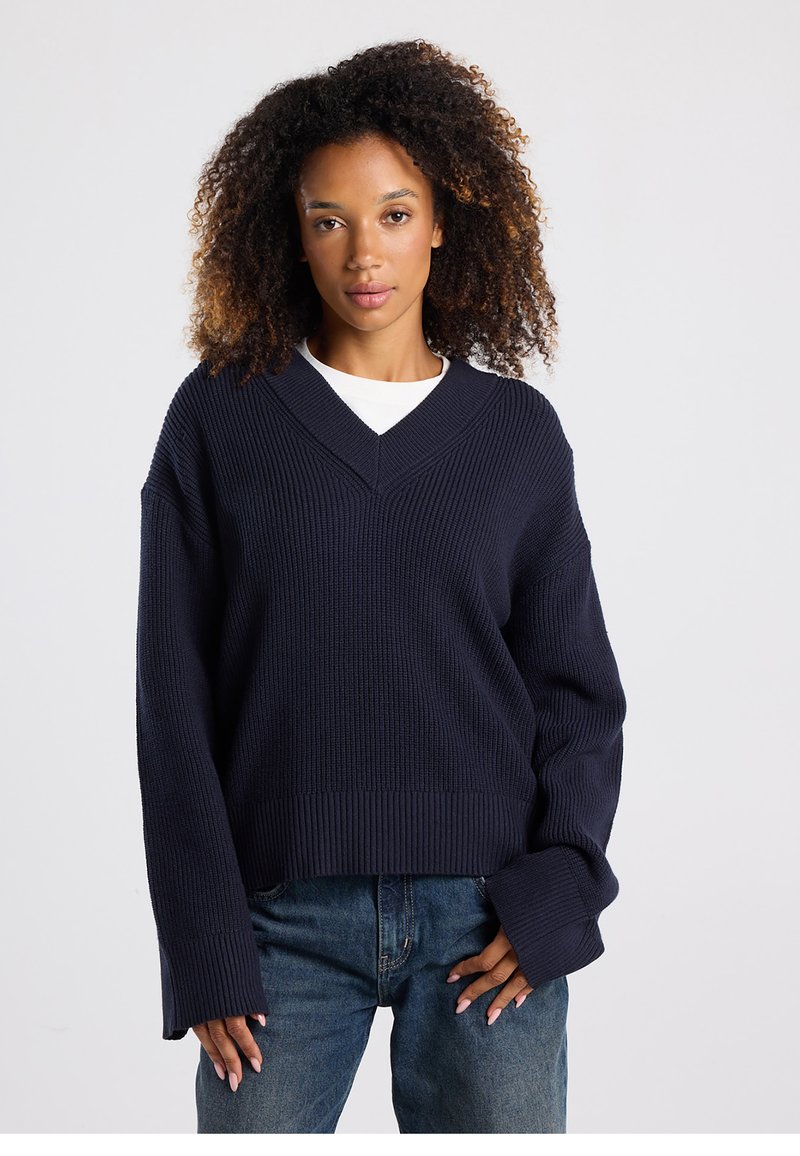 Navy blue oversized sweater with a V-neck, ribbed texture, dropped shoulders, and long sleeves, paired with denim jeans.
