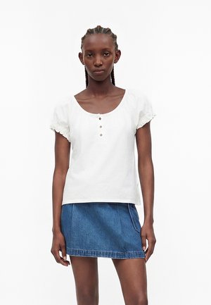 Noor naine punutud juustega, kes kannab valget lühikeste varrukatega nööbitavat pluusi ja sinist teksast miniseelikut, seisab valge tausta ees.