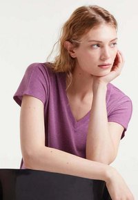 Joven mujer con cabello claro que lleva una camiseta morada con cuello en V, apoyando su chin en la mano, mirando pensativa hacia un lado.