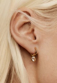 Pendiente de aro dorado con una gema ovalada clara colgante. Primer plano del pendiente sobre un fondo de tono de piel claro y cabello rubio.