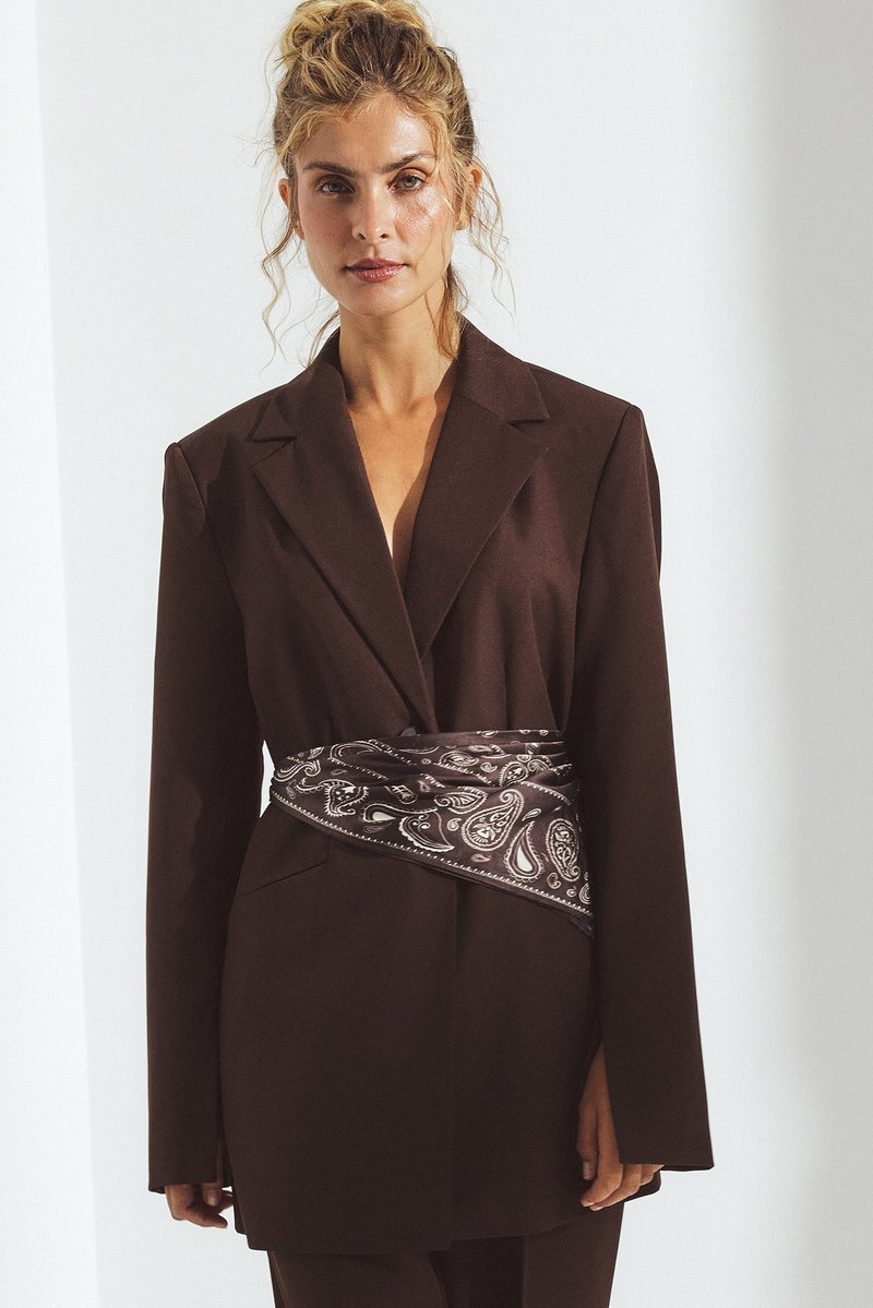 Brūns pielāgots žakets ar garām piedurknēm, ar rakstu tumši brūnā un baltā šallē, kas piesaistīta pie vidukļa. Līdzena auduma un strukturēta forma.