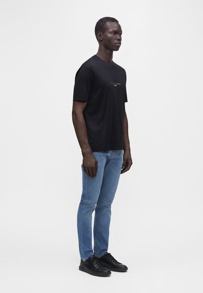 Άνδρας που στέκεται, φοράει μαύρο T-shirt με λευκό κείμενο, μπλε τζιν και μαύρα αθλητικά παπούτσια μπροστά σε απλό φωτεινό φόντο.